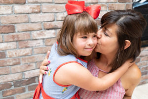 Mom kissing her daughters cheek who is wearing a red ribbon in her hair