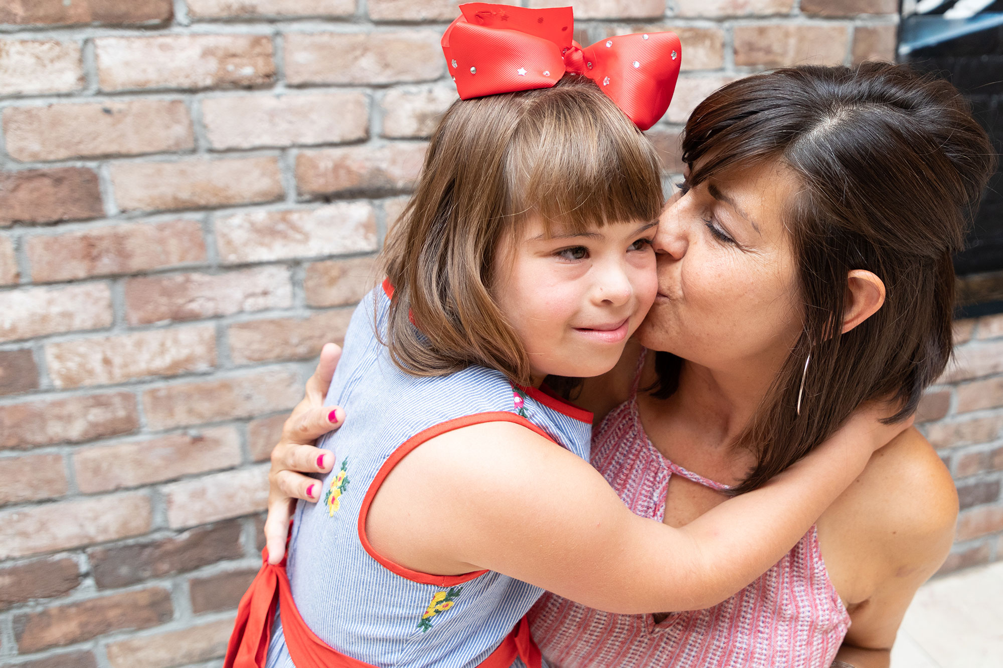 Mom kissing her daughters cheek who is wearing a red ribbon in her hair