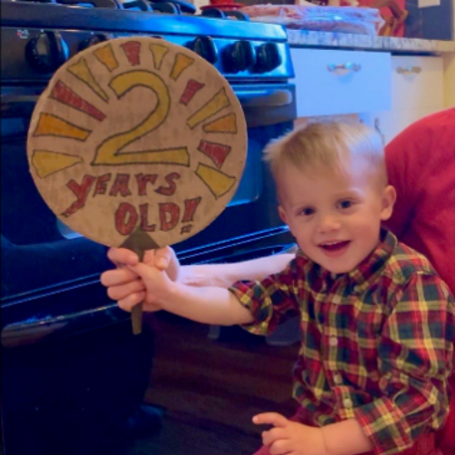 A little boy smiling holding up a round sign that says 2 years old!