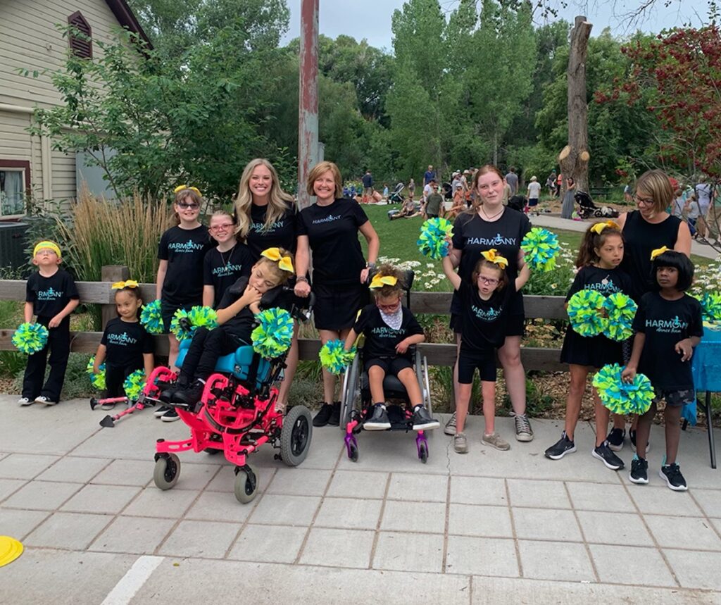 A group of adults and children are outside in black uniforms that read "Harmony" and holding pom-poms. Some of the children are in wheelchairs.