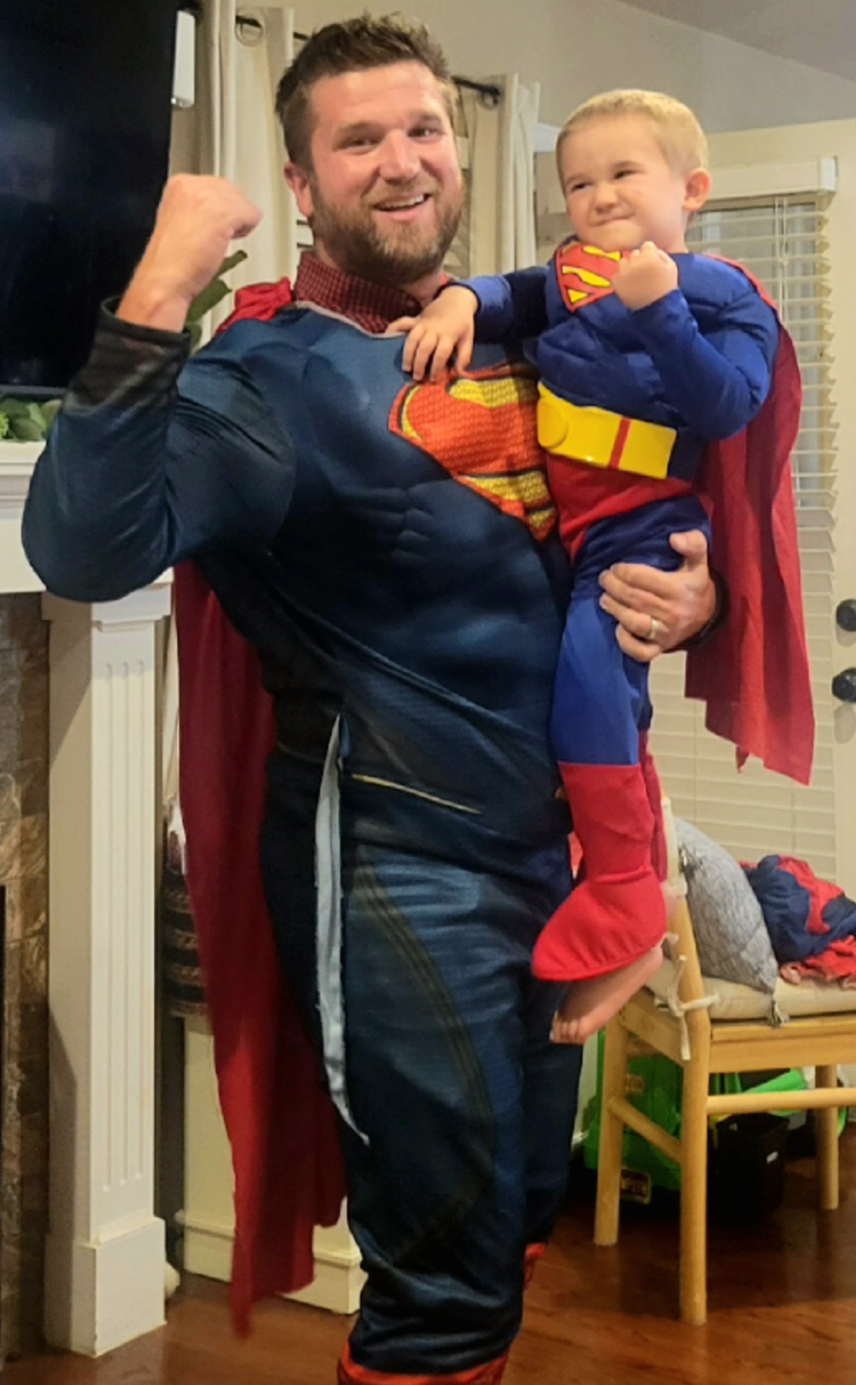 One adult holding a child. Both have a raised fist and are wearing blue outfits, red capes, and have a large red "S" in a yellow diamond on chests.