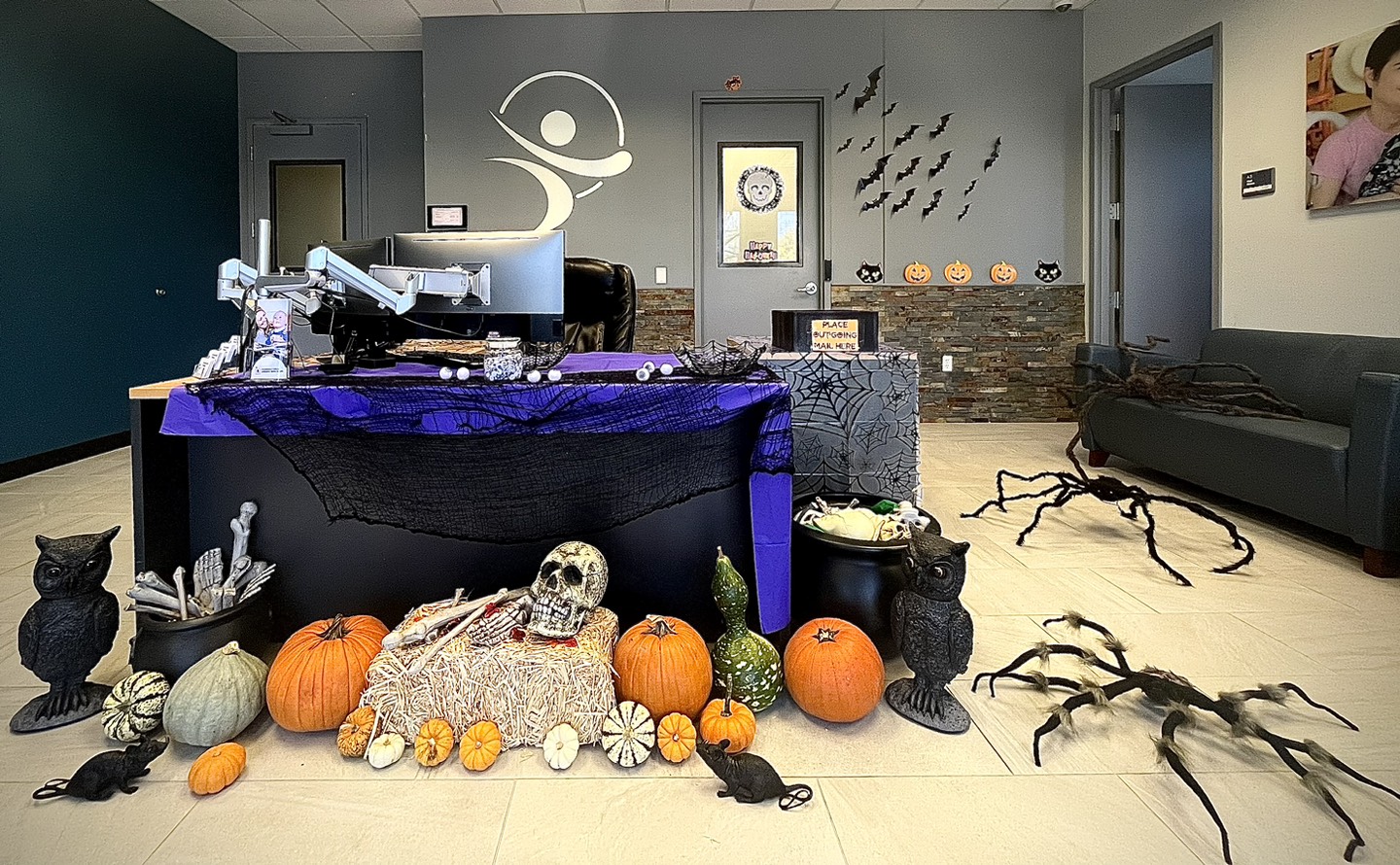 Office area with large spiders on the ground, black spiderwebs draped on the desks, pumpkins of different sizes in front of desk, with a skull and bones in cauldrons, black owls and rats, and bats on the wall.