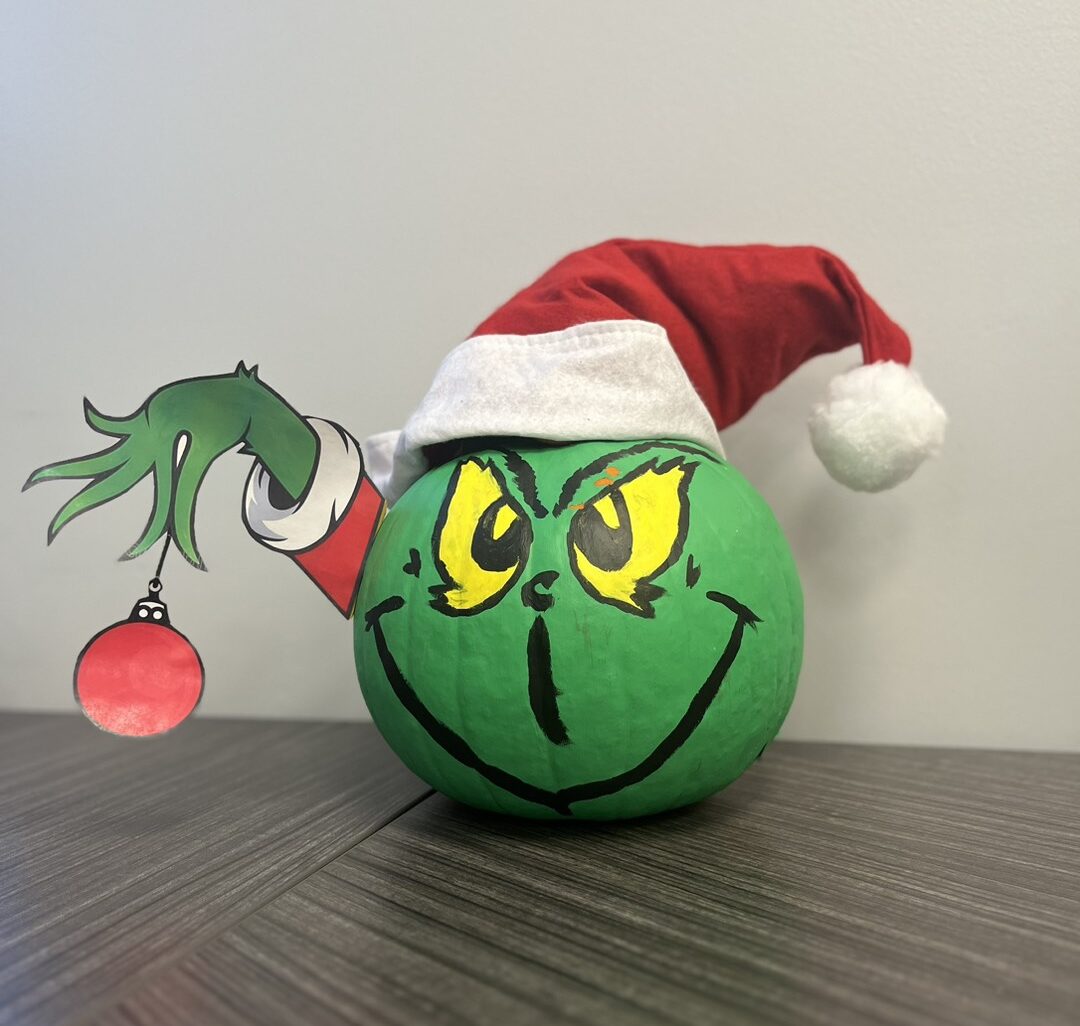 Green painted pumpkin with yellow eyes and face drawn on in black. It has a red and white pointed hat with ball on end. It has a printed out green arm with a red and white sleeve holding a red circle ornament.
