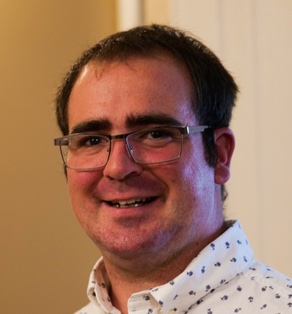 A portrait of a man with dark brown hair and glasses smiling at the camera.