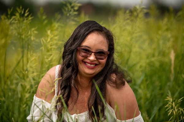 A woman in sunglasses sat down on the ground surrounded by tall grass, looking at the camera and smiling.