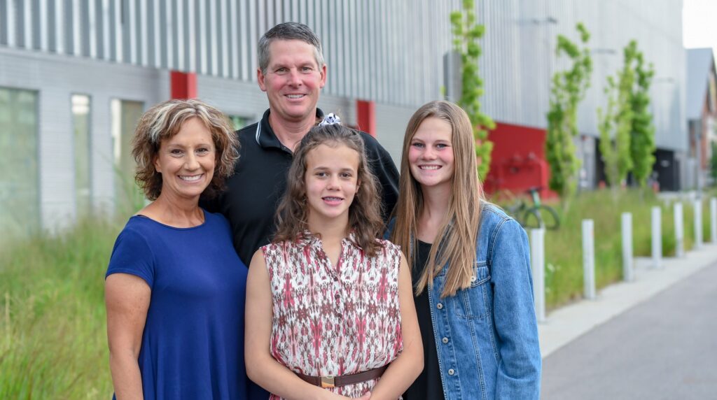 A family smiles at the camera while standing outside. A mother and father are on the left and two teen daughters are on the right.