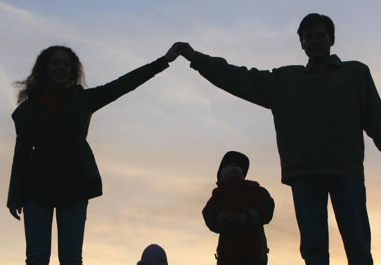 A silhouette of a family standing in front of the sky. The mother and father hold hands in the air, while a child looks up underneath them.