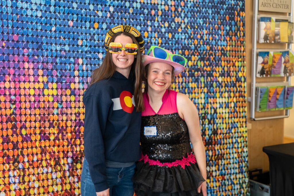 2 females in our community are smiling wearing funny hats and sunglasses standing in front of a multicolored screen