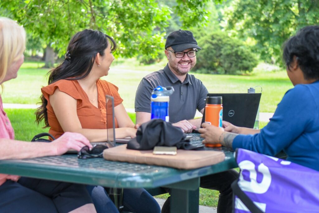 Four team members sitting at a picnic table in the park looking at the male employee in glasses smiling