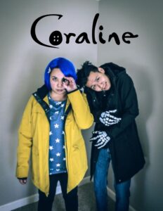 Two adults one in a blue wig, yellow raincoat, and blue and white stars shirt holding a button to their eye. The other person is hunched over wearing a black coat and skeleton gloves.