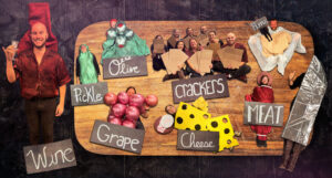 Team placed on a wooden charcuterie board dressed as different objects labeled as wine, pickle, grape, olive, crackers, cheese, meat, hummus and a cheese knife.