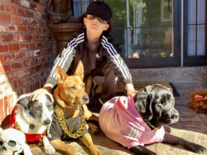 Person with black bucket hat, sunglasses and black track jacket with white stripes posing with three dogs in different color hoodies - red, black, and pink.