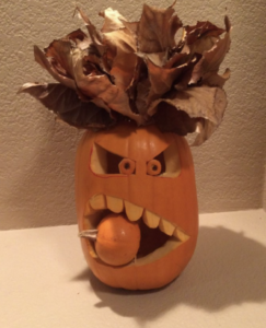 Pumpkin carving with leaves at the top to look like hair. The face looks like it is sneering with a smaller pumpkin held in between its teeth.