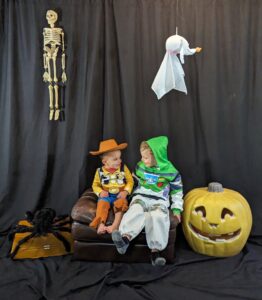 Two young children sitting in-between a spider and jack o lantern. One on the left wearing cowboy/woody from toy story costume, other one on the right wearing space suit/buzz lightyear costume.
