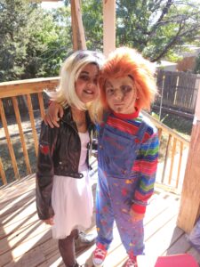 Two children - one in black leather jacket, white dress, and blonde wig, while other is in overalls. multicolored striped shirt, and red wig.
