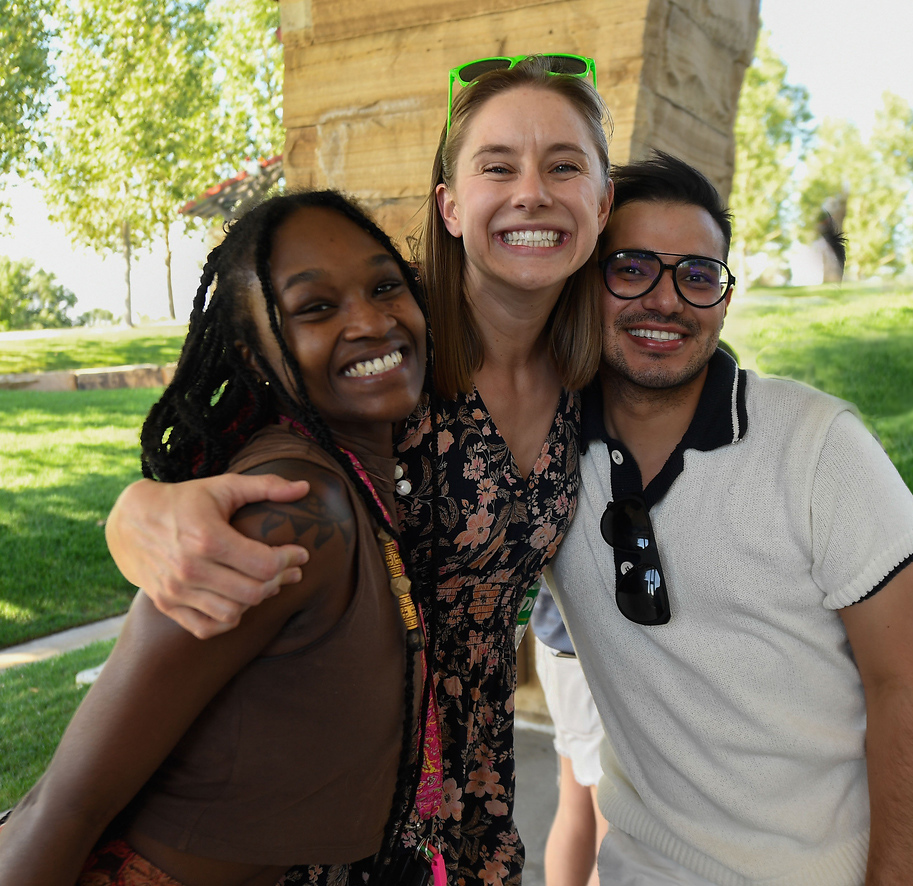 Three team members smiling with on team member with green sunglasses on hugging the other two