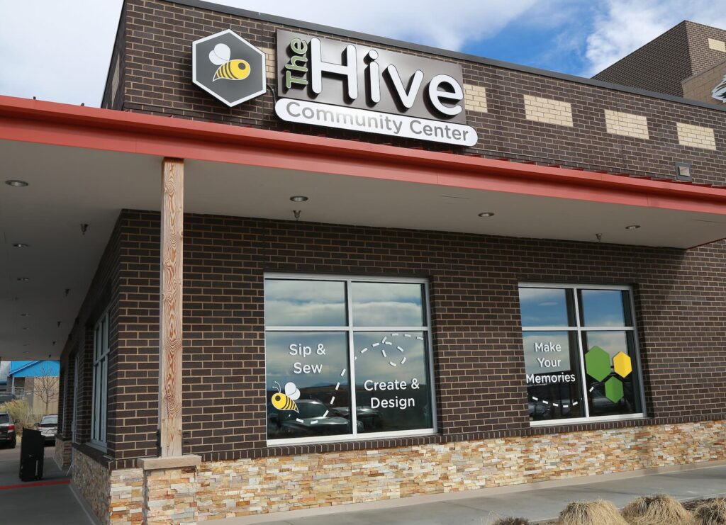A photo of an exterior of a building. The sign on the building reads "The Hive Community Center" and has a logo of a bee. The windows read "Sip & Sew. Create & Design. Make your Memories."