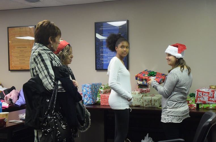 Woman in a Santa hat handing out a gift to a mother and daughter and a teen looking at the camera