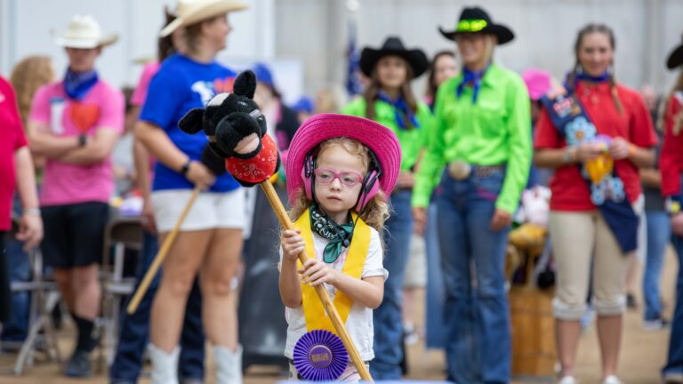 A small child wearing a bright pink cowboy hat, pink glasses, and over-ear headphones stands indoors holding a stick with a small plush horse head on top. A yellow ribbon with a purple rosette hangs around the child’s neck. In the background, several people dressed in colorful western-style clothing and cowboy hats are gathered, slightly out of focus.